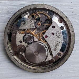 May include: Close-up of a vintage watch movement, showcasing intricate gears, springs, and other mechanical components. The metal parts are silver and gold, with a circular design. The watch movement is encased in a silver frame. The text "2409A" is visible.