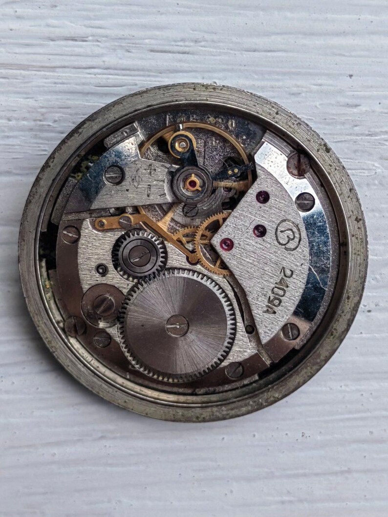 May include: Close-up of a vintage watch movement, showcasing intricate gears, springs, and other mechanical components. The metal parts are silver and gold, with a circular design. The watch movement is encased in a silver frame. The text "2409A" is visible.