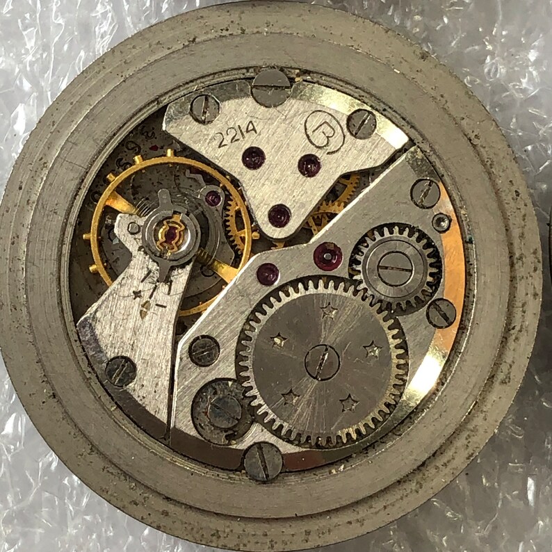 May include: Close-up of a silver watch movement with gold gears and a black dial. The movement has a number "2214" and a letter "B" printed on it.