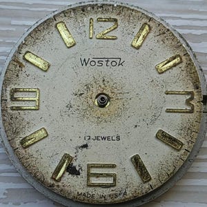K&ouml;nnte beinhalten: Vintage Wostok Zifferblatt mit cremefarbenem Zifferblatt und goldfarbenen Stundenmarkierungen. Das Zifferblatt zeigt die Zahlen 12, 3, 6 und 9. Der Text "Wostok" und "17 JEWELS" sind sichtbar. Die Worte "MADE IN USSR" sind ebenfalls sichtbar.