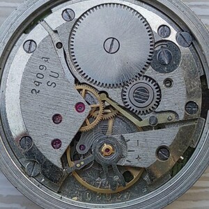 May include: Close-up of a watch movement with gears, screws, and other mechanical components. The metal parts are silver and gray, with some gold-colored elements. The text "2409A SU" is visible on a metal plate.