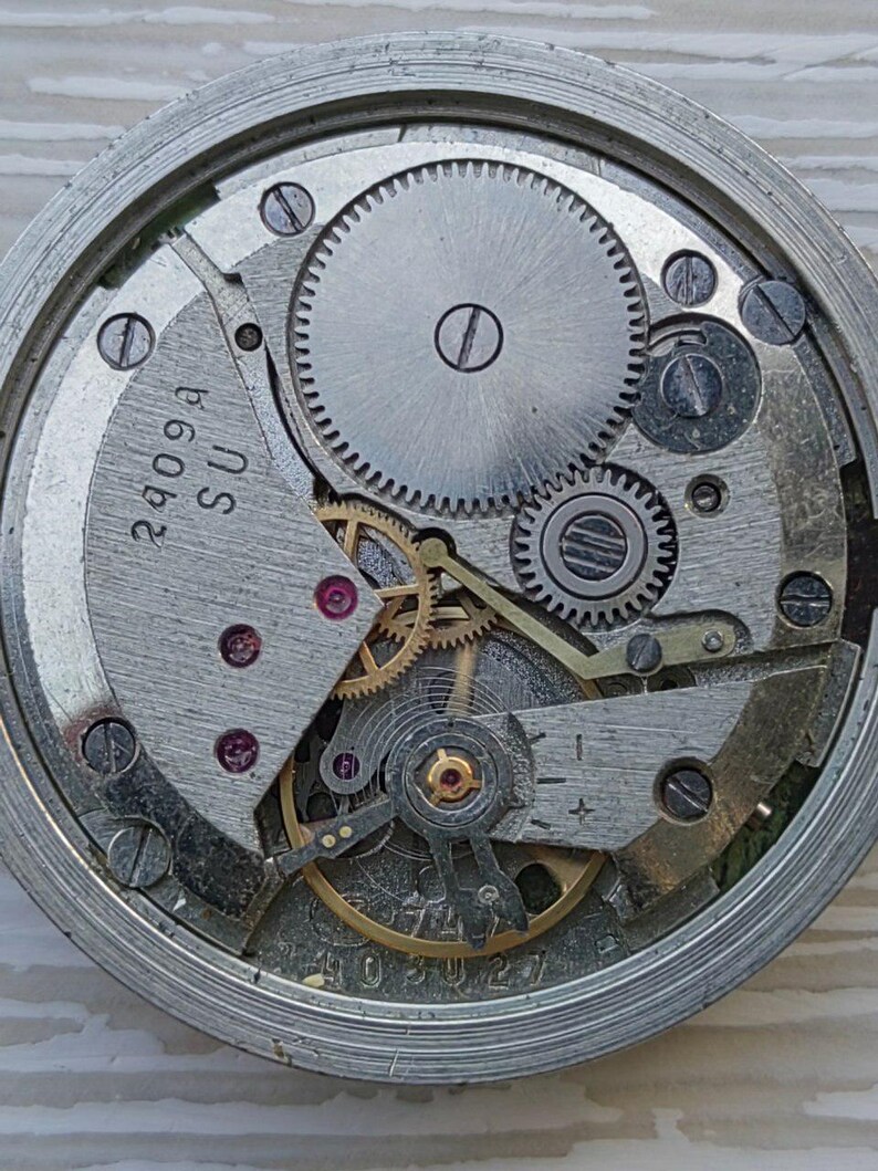 May include: Close-up of a watch movement with gears, screws, and other mechanical components. The metal parts are silver and gray, with some gold-colored elements. The text "2409A SU" is visible on a metal plate.