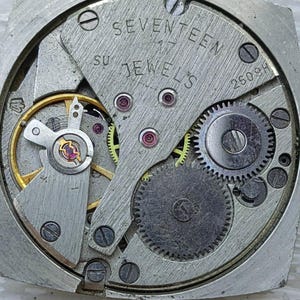 K&ouml;nnte beinhalten: Nahaufnahme eines Vintage-Uhrwerks. Der silberfarbene Mechanismus verf&uuml;gt &uuml;ber Zahnr&auml;der, Juwelen und den Text "SEVENTEEN 17 JEWELS". Das Uhrwerk befindet sich in einem quadratischen Geh&auml;use.