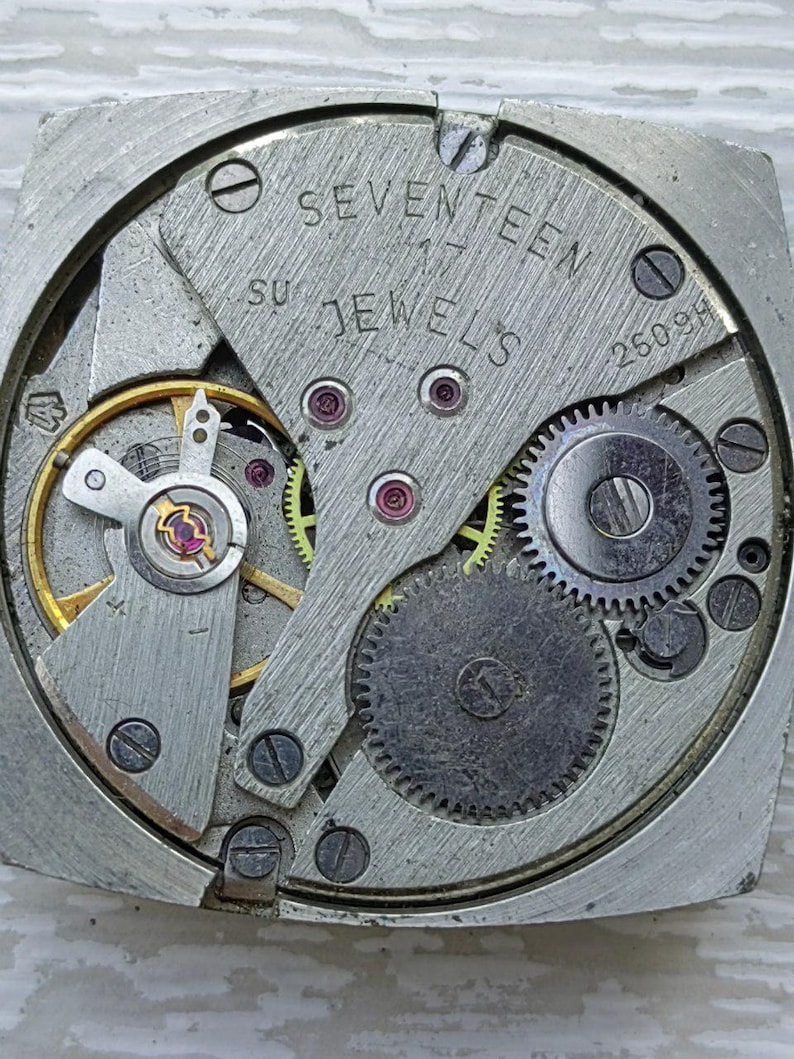 K&ouml;nnte beinhalten: Nahaufnahme eines Vintage-Uhrwerks. Der silberfarbene Mechanismus verf&uuml;gt &uuml;ber Zahnr&auml;der, Juwelen und den Text "SEVENTEEN 17 JEWELS". Das Uhrwerk befindet sich in einem quadratischen Geh&auml;use.