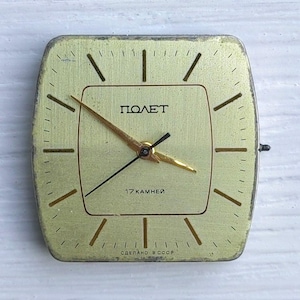 May include: A vintage square watch face with a gold-colored face and hands. The watch face has the word "ПОЛЕТ" and the text "17 KAMHEЙ" printed on it. The watch face is approximately 1.5 inches square.