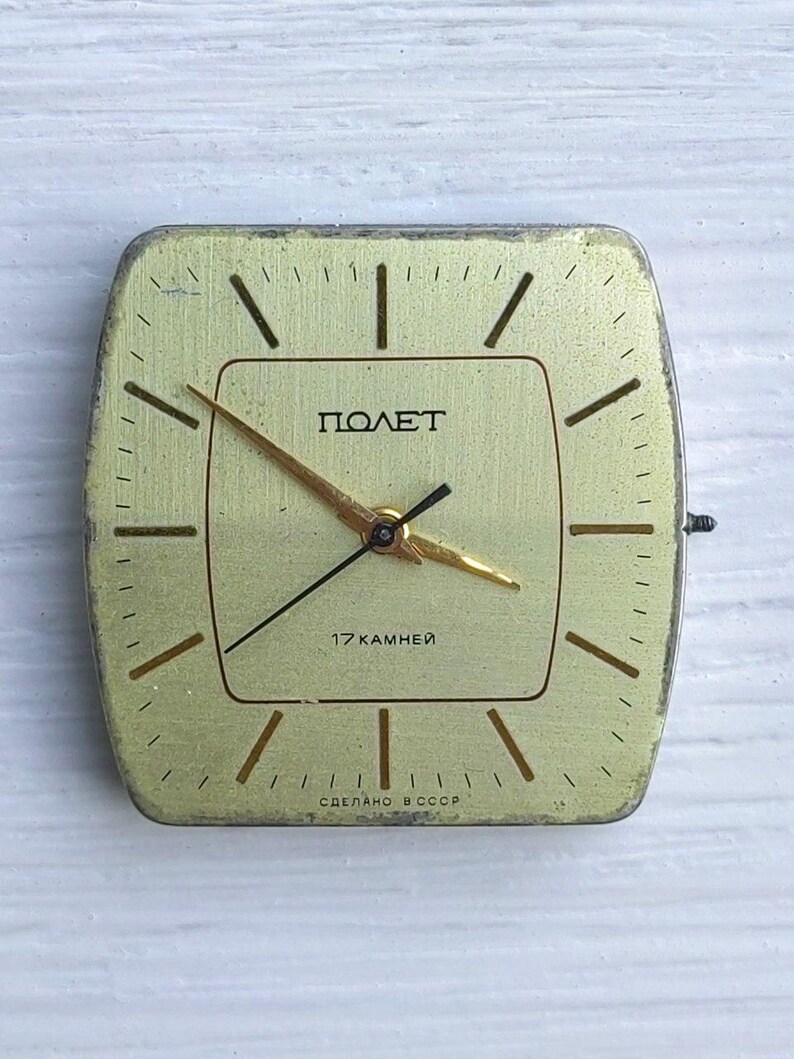 May include: A vintage square watch face with a gold-colored face and hands. The watch face has the word "ПОЛЕТ" and the text "17 KAMHEЙ" printed on it. The watch face is approximately 1.5 inches square.