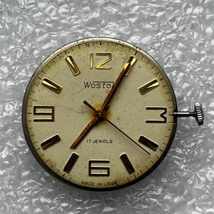 May include: Vintage Wostok watch face with a cream-colored dial and gold-tone hands and markers. The watch face features the number 12, 3, 6, and 9 in gold. The text "Wostok" and "17 Jewels" are visible. The watch face is marked "Made in USSR".