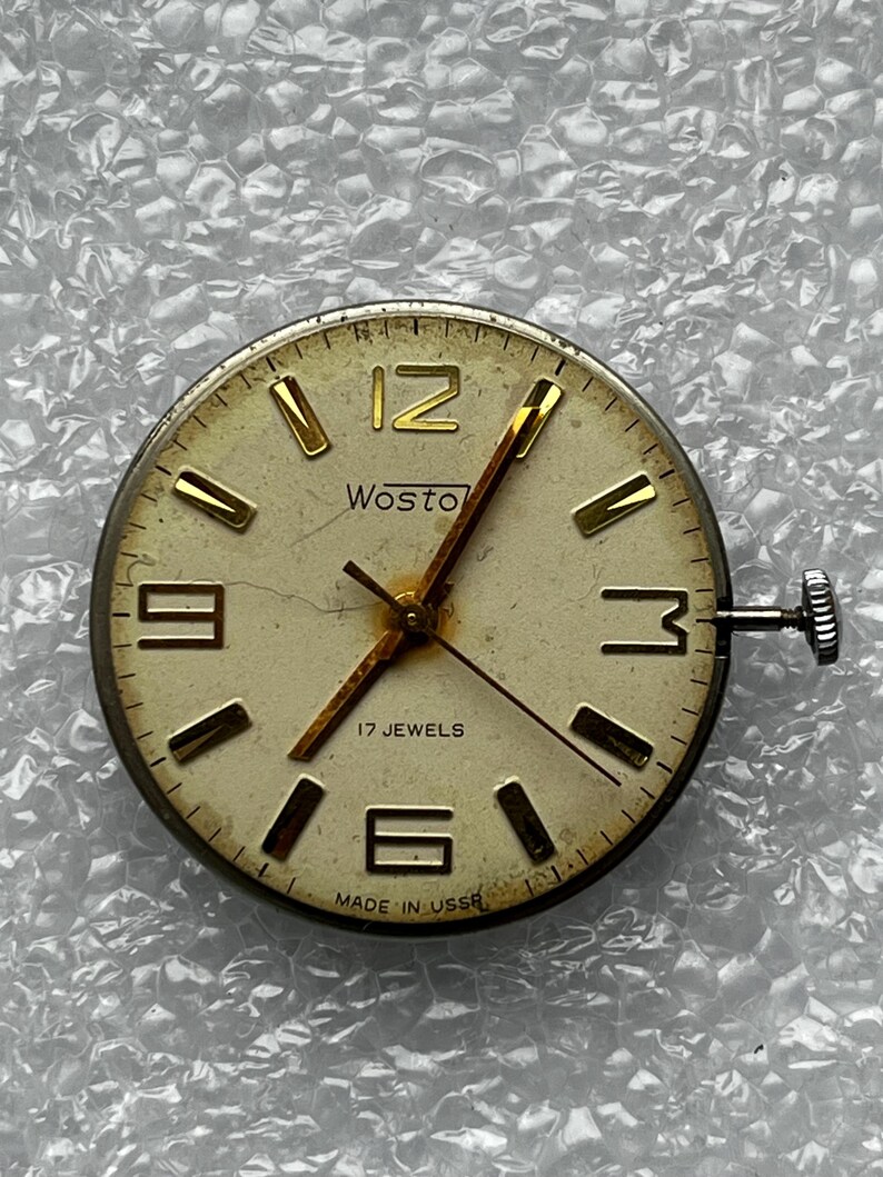 May include: Vintage Wostok watch face with a cream-colored dial and gold-tone hands and markers. The watch face features the number 12, 3, 6, and 9 in gold. The text "Wostok" and "17 Jewels" are visible. The watch face is marked "Made in USSR".