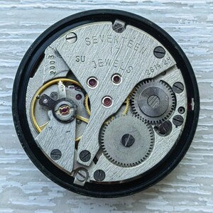 May include: Close-up of a watch movement with visible gears, screws, and the text "SEVENTEEN 17 JEWELS". The movement is housed in a black circular frame. The image shows intricate mechanical details.
