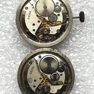 May include: Two vintage watch movements with exposed gears and intricate mechanical components. The silver-toned metal movements feature the text "2409A" and a crown. The gears are visible, showcasing the inner workings of the timepieces.