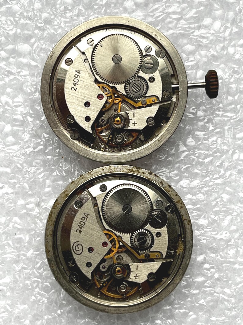 May include: Two vintage watch movements with exposed gears and intricate mechanical components. The silver-toned metal movements feature the text "2409A" and a crown. The gears are visible, showcasing the inner workings of the timepieces.
