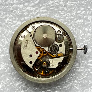 May include: Close-up of a watch movement with visible gears, springs, and a winding stem. The metal components are silver-toned, with some gold-colored gears. The text "2409A" is visible on a white plate.