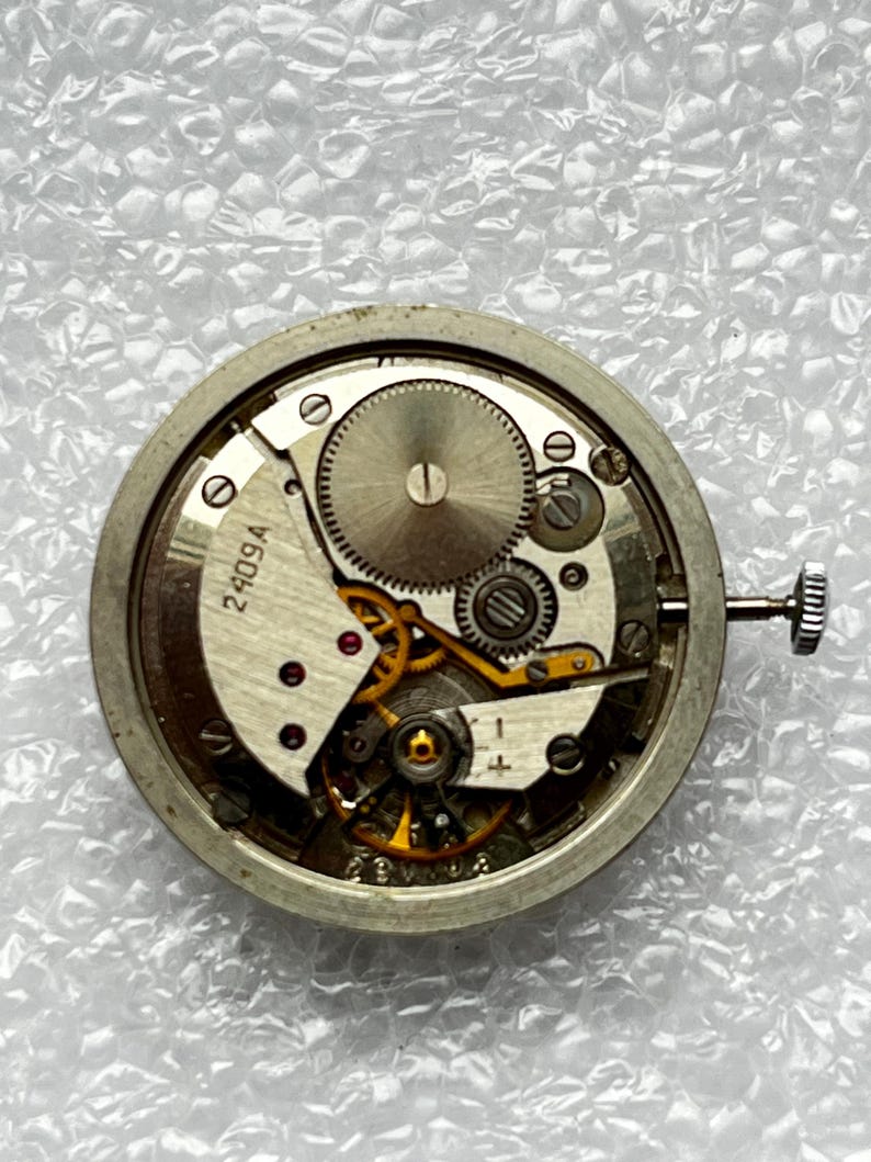 May include: Close-up of a watch movement with visible gears, springs, and a winding stem. The metal components are silver-toned, with some gold-colored gears. The text "2409A" is visible on a white plate.