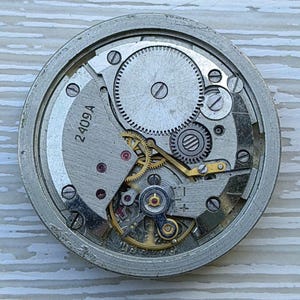 K&ouml;nnte beinhalten: Nahaufnahme eines Vintage-Uhrwerks mit sichtbaren Zahnr&auml;dern, Federn und der Nummer "2409A" auf einer silberfarbenen Platte. Die komplizierten mechanischen Komponenten sind in einem kreisf&ouml;rmigen Rahmen angeordnet und zeigen das Innenleben einer Uhr.