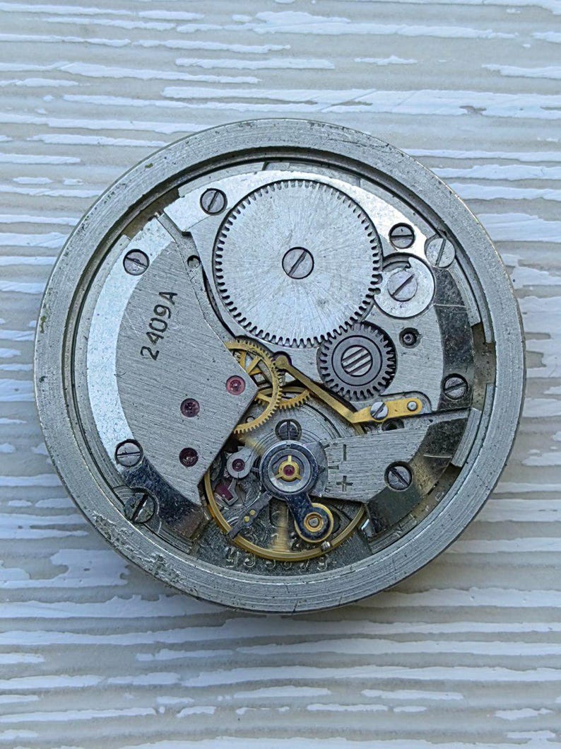 K&ouml;nnte beinhalten: Nahaufnahme eines Vintage-Uhrwerks mit sichtbaren Zahnr&auml;dern, Federn und der Nummer "2409A" auf einer silberfarbenen Platte. Die komplizierten mechanischen Komponenten sind in einem kreisf&ouml;rmigen Rahmen angeordnet und zeigen das Innenleben einer Uhr.