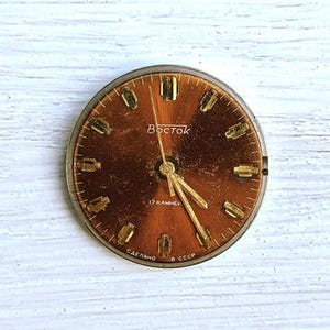 May include: A vintage watch face with a brown dial and gold-colored hands and hour markers. The word "Восток" is printed in Cyrillic script. The watch face also has the text "17 камней" and "СДЕЛАНО В СССР".
