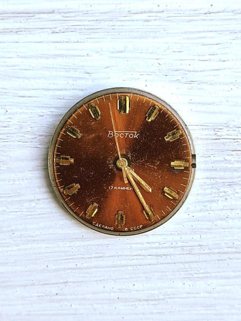 May include: A vintage watch face with a brown dial and gold-colored hands and hour markers. The word "Восток" is printed in Cyrillic script. The watch face also has the text "17 камней" and "СДЕЛАНО В СССР".