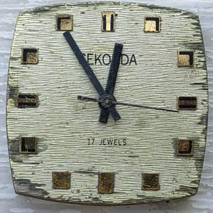 K&ouml;nnte beinhalten: Vintage Eko Da Armbanduhr mit strukturiertem, cremefarbenem Zifferblatt und goldfarbenen quadratischen Markierungen. Die Uhr hat schwarze Zeiger und den Text "17 JEWELS". Das Zifferblatt ist quadratisch mit abgerundeten Kanten.