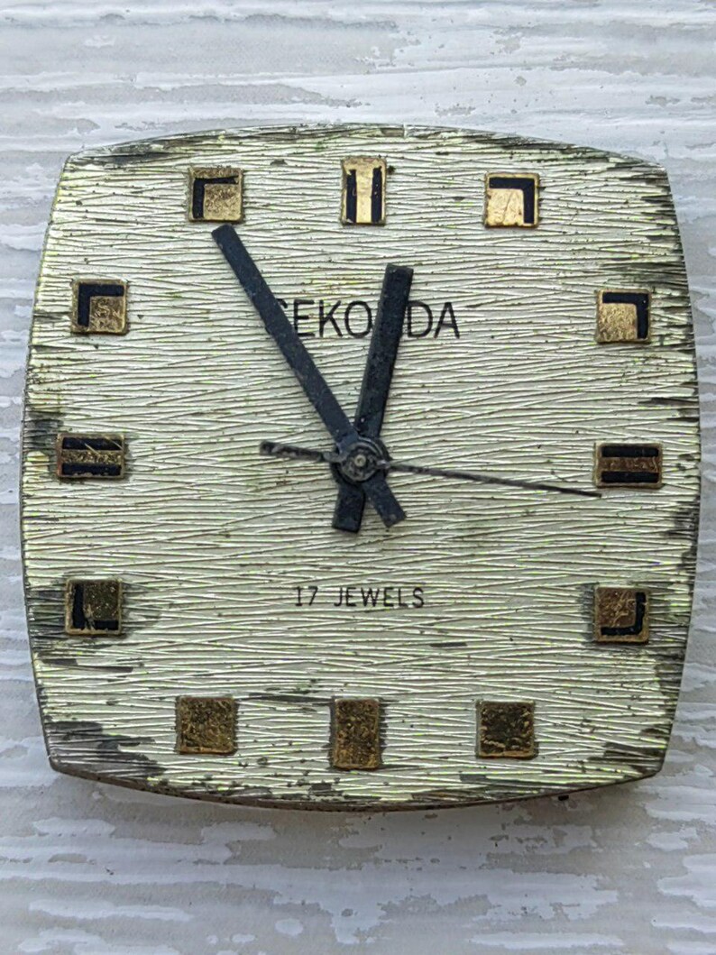 K&ouml;nnte beinhalten: Vintage Eko Da Armbanduhr mit strukturiertem, cremefarbenem Zifferblatt und goldfarbenen quadratischen Markierungen. Die Uhr hat schwarze Zeiger und den Text "17 JEWELS". Das Zifferblatt ist quadratisch mit abgerundeten Kanten.