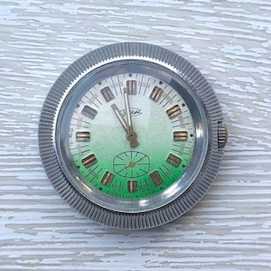 Puede incluir: Un reloj de pulsera vintage con un bisel plateado y acanalado. La esfera del reloj tiene un degradado verde, con marcadores de hora y agujas rectangulares doradas. La esfera del reloj también tiene el texto "GUST" y "Сделано en la URSS".
