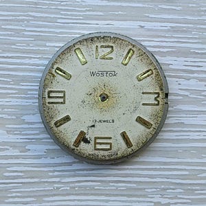 K&ouml;nnte beinhalten: Vintage Wostok Zifferblatt mit cremefarbenem Zifferblatt und goldfarbenen Ziffern. Das Zifferblatt tr&auml;gt den Markennamen "Wostok", "17 Jewels" und "Made in USA". Das Zifferblatt weist Gebrauchsspuren und Alterungserscheinungen auf.