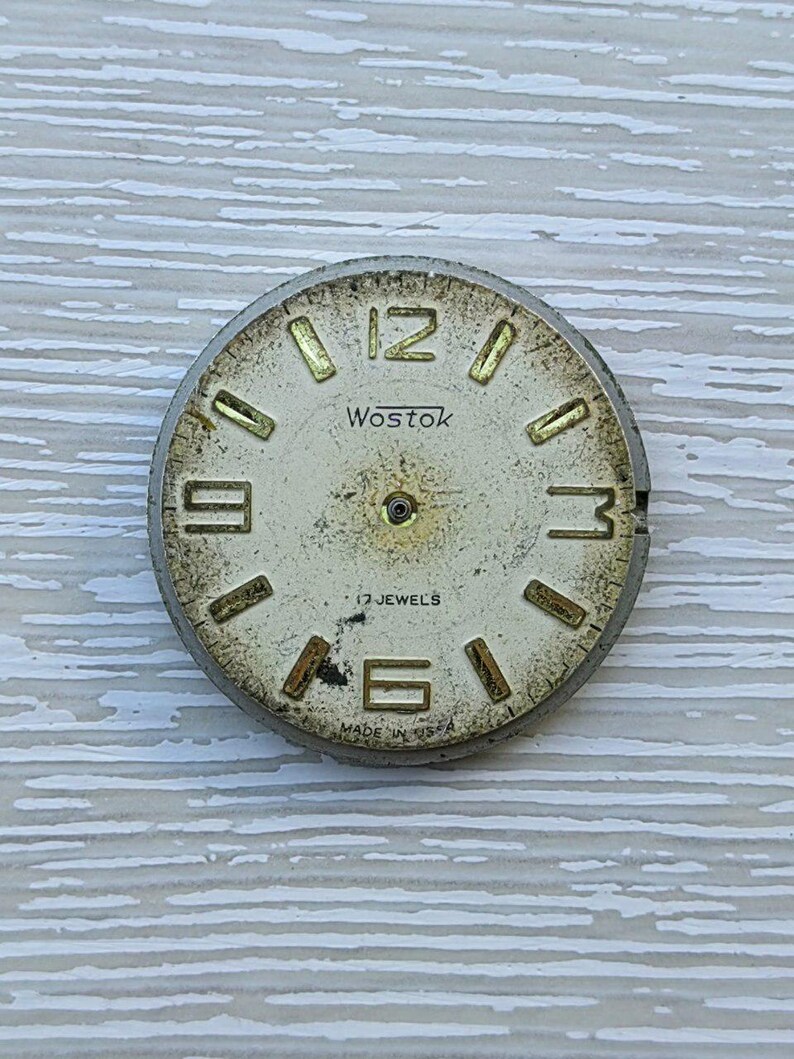 K&ouml;nnte beinhalten: Vintage Wostok Zifferblatt mit cremefarbenem Zifferblatt und goldfarbenen Ziffern. Das Zifferblatt tr&auml;gt den Markennamen "Wostok", "17 Jewels" und "Made in USA". Das Zifferblatt weist Gebrauchsspuren und Alterungserscheinungen auf.
