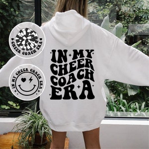 May include: White hoodie with the text "IN MY CHEER COACH ERA" in black, retro-style lettering on the back. Two circular graphics on the front feature a pom-pom and a smiley face with the same text. A casual, comfortable garment.