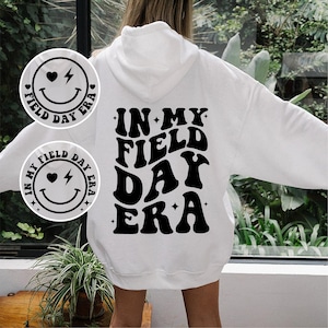 May include: White hoodie with a black graphic that says "In My Field Day Era" in a retro font. The graphic is surrounded by small stars. There is a smaller black and white graphic of a smiley face with a heart and lightning bolt on the hoodie.