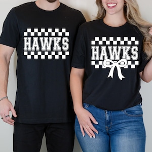May include: Black t-shirts with the word "HAWKS" in white block letters, framed by a checkered pattern. One shirt has a bow graphic below the text. The shirts are worn by two people.