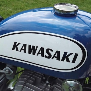 Può includere: Primo piano del serbatoio di una moto d'epoca. Il serbatoio è blu lucido con una grafica ovale bianca con la scritta "KAWASAKI" in nero. È visibile un tappo del carburante cromato.