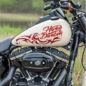 Puede incluir: Tanque de gasolina de motocicleta color crema con las palabras "Harley Davidson" en escritura roja y diseños tribales rojos. La motocicleta tiene componentes negros y está sobre un fondo verde borroso.
