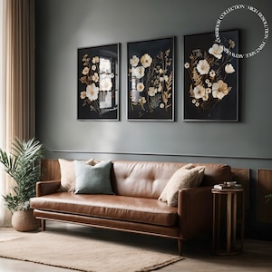 Puede incluir: Tres impresiones enmarcadas de flores blancas sobre un fondo oscuro, colgadas en una pared gris sobre un sofá de cuero marrón con almohadas.