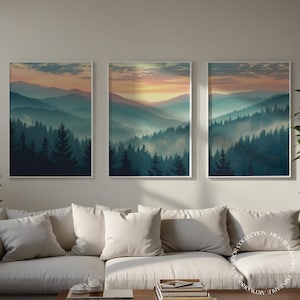 Może przedstawiać: Zestaw trzech oprawionych wydruków przedstawiających mglisty krajobraz górski ze wschodem słońca nad lasem. Wydruki są w białej ramie i mają stonowaną paletę kolorów w odcieniach niebieskiego, zielonego i pomarańczowego.