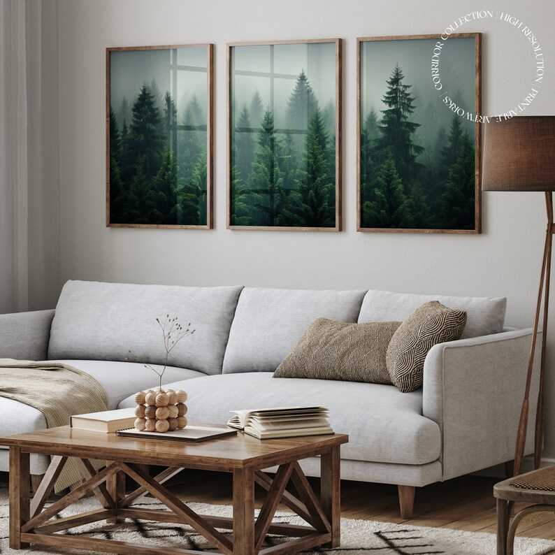 K&ouml;nnte beinhalten: Drei gerahmte Drucke einer nebeligen Waldszene mit hohen immergr&uuml;nen B&auml;umen. Die Drucke h&auml;ngen an einer wei&szlig;en Wand &uuml;ber einem hellgrauen Ecksofa mit einer braunen Wolldecke und einem Holz-Couchtisch.
