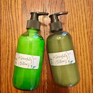 Könnte beinhalten: Zwei Glasflaschen mit schwarzen Pumpspendern, gefüllt mit grüner Flüssigkeit. Jede Flasche hat ein weißes Etikett mit dem Text "Graceful Tallow". Die Flaschen stehen auf einer Holzoberfläche.