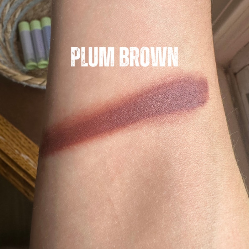 K&ouml;nnte beinhalten: Ein Lippenstift-Swatch in Pflaumenbraun wird auf einem Arm gezeigt. Der Text "PLUM BROWN" ist in wei&szlig;er, fetter Schrift &uuml;berlagert. Die Lippenstiftfarbe ist ein tiefes, r&ouml;tlich-braunes.