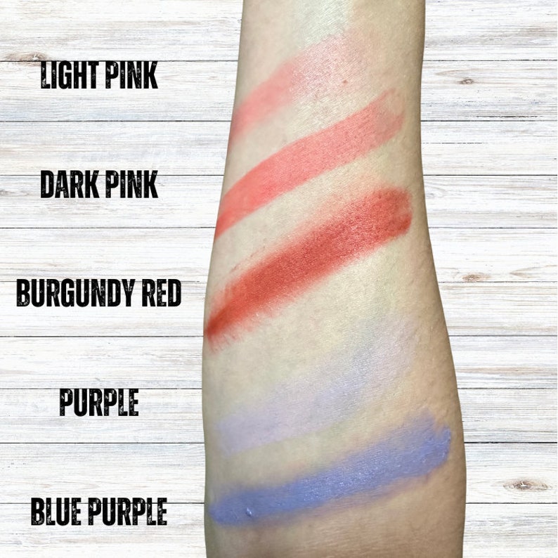K&ouml;nnte beinhalten: Lippenstift-Farbmuster auf einem Arm. Die Farben sind hellrosa, dunkelrosa, burgunderrot, lila und blau-lila. Die Namen der Farben sind in schwarzen Gro&szlig;buchstaben &uuml;ber jedem Muster geschrieben.