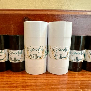 Op de afbeelding: Een verzameling cilindervormige containers, zwart en wit, staat op een houten oppervlak. Elke container is voorzien van een etiket met "Graceful Tallow" in sierlijke letters, met tarwe- en bladmotieven.