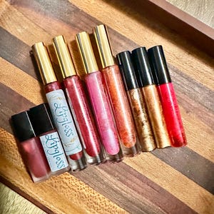 May include: A selection of lip gloss tubes and small containers in shades of pink, red, and gold. The tubes have gold caps and the word "Lipgloss" is printed on the labels. The items are arranged on a wooden tray with a striped pattern.