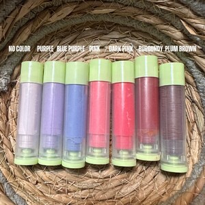 K&ouml;nnte beinhalten: Eine Auswahl an Lippenbalsam in verschiedenen Farben, darunter Lila, Rosa und Braun, mit gr&uuml;nen Kappen. Die Lippenbalsame sind auf einem geflochtenen Korb angeordnet, mit Farbetiketten dar&uuml;ber.