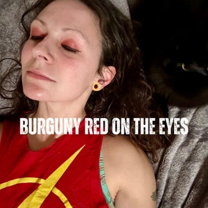 K&ouml;nnte beinhalten: Eine Person mit burgunderfarbenem Lidschatten und einem burgunderfarbenen Tanktop mit gelbem Design. Der Text "BURGUNY RED ON THE EYES" wird in Wei&szlig; &uuml;ber dem Bild angezeigt. Im Hintergrund ist eine schwarze Katze zu sehen.