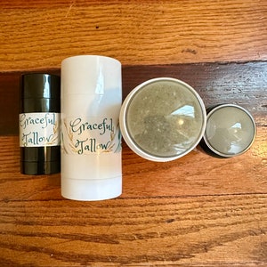 Op de afbeelding: Drie deodorant sticks, twee zijn wit met een groen en gouden label met de tekst "Graceful Tallow" en een is zwart met hetzelfde label. De sticks liggen op een houten oppervlak.