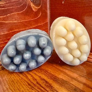 May include: Two oval soaps with textured surfaces. One is light blue, the other off-white. Each soap has rounded protrusions. The soaps are displayed on a wooden surface with a reddish-brown hue.