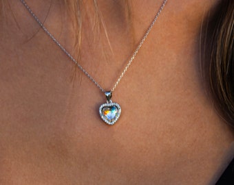 May include: A silver necklace with a heart-shaped pendant. The pendant has a shimmering, iridescent center and is surrounded by small, clear crystals. The necklace is displayed against a dark background.