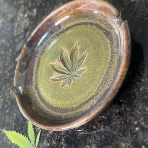 May include: Oval ceramic ashtray with a cannabis leaf design. The ashtray has a green interior with a raised leaf and a brown rim. It features a small notch for resting items.
