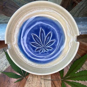 May include: A ceramic ashtray with a blue and tan glaze. The center features a raised cannabis leaf design. The ashtray has two small notches for holding items. The ashtray is round and sits on a wooden surface.