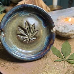 May include: A ceramic ashtray with a cannabis leaf design. The ashtray is round with a raised leaf in the center and a dark blue and brown glaze. A dried cannabis leaf rests on a gold surface.