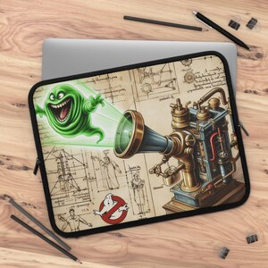 May include: Laptop sleeve featuring a green cartoon ghost emitting a beam towards a detailed steampunk-style device. The design includes blueprints, the Ghostbusters logo, and a black border.