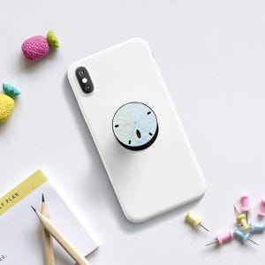 May include: A white phone case with a round, light blue and white sand dollar design phone grip. The phone case is on a white surface with colorful push pins, pencils, and decorative erasers.