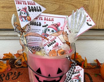 May include: A pink Halloween bucket with a jack-o'-lantern face design, filled with treats and decorations. The bucket includes a flag and tags that read "You've Been Booed", a skeleton hand, and a small pumpkin. The bucket is on a doormat.